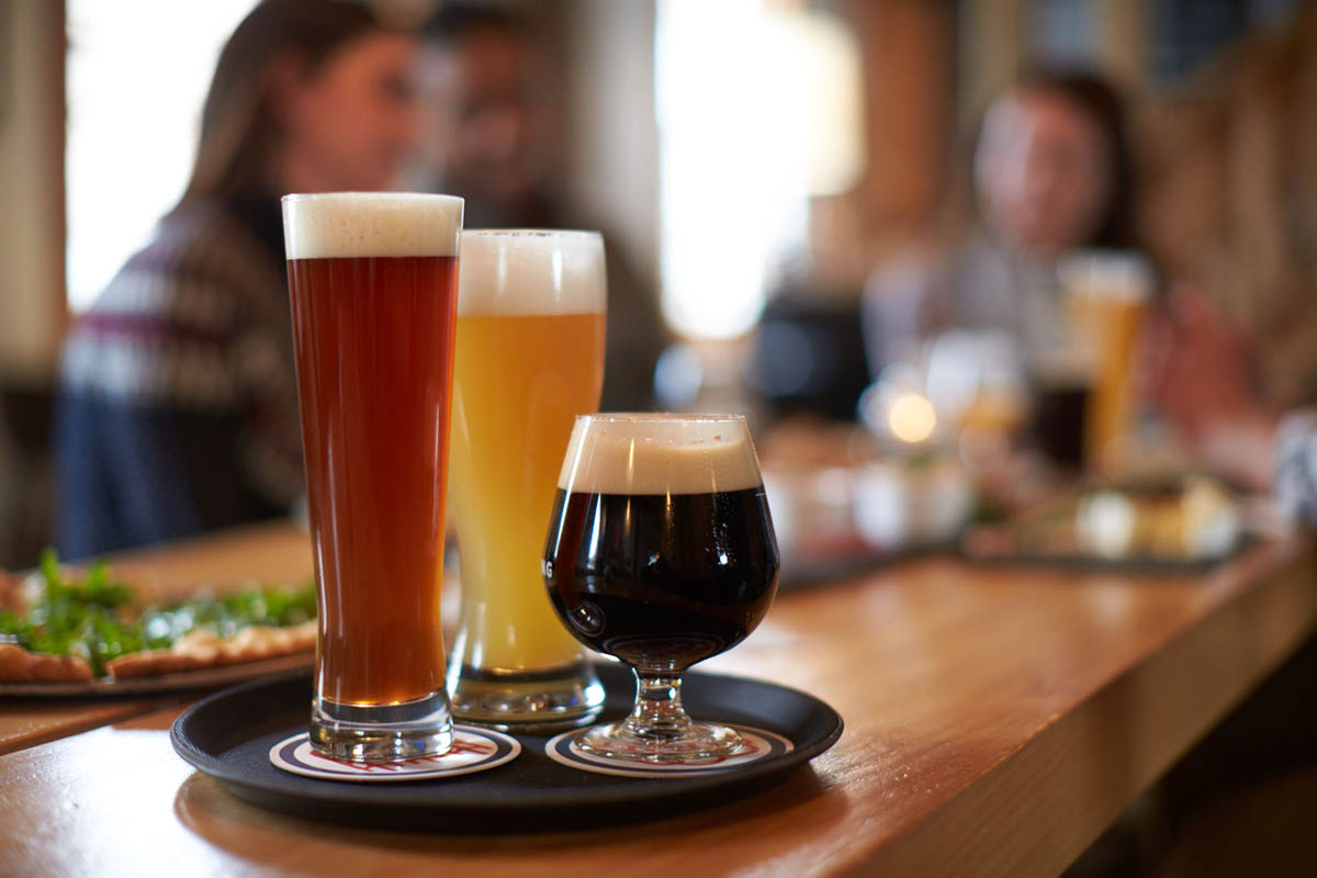 three different color beers in three different shape glasses sitting on a tray on a bar with an out of focus background